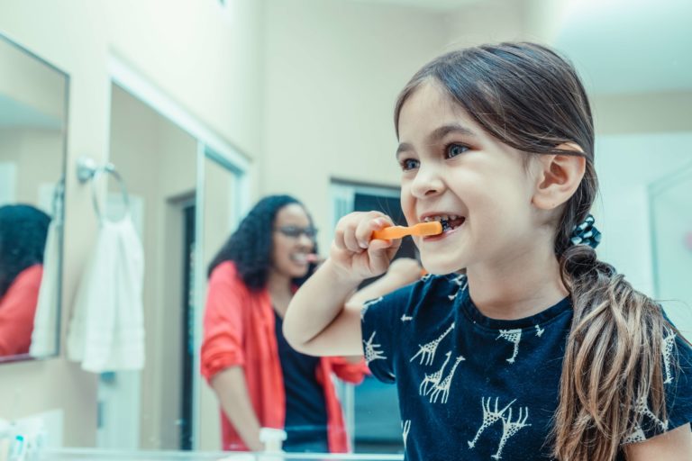 Young girl brushes her teeth happily while her mother looks on approvingly.